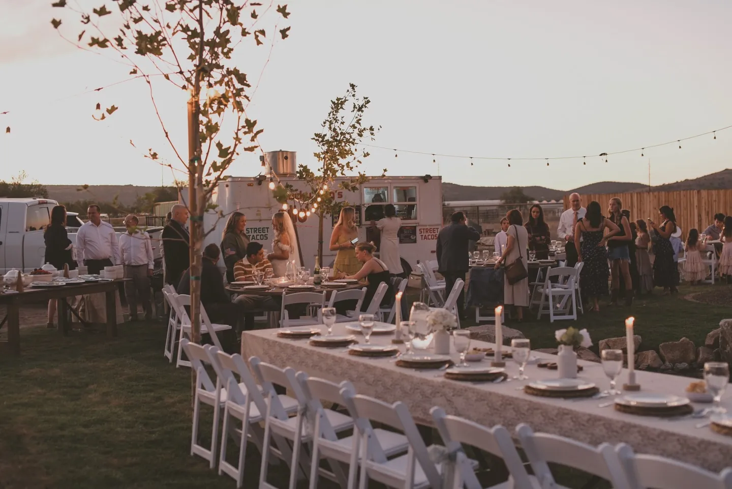 A long dinner table at dusk with place settings, candles and guests gathering around it