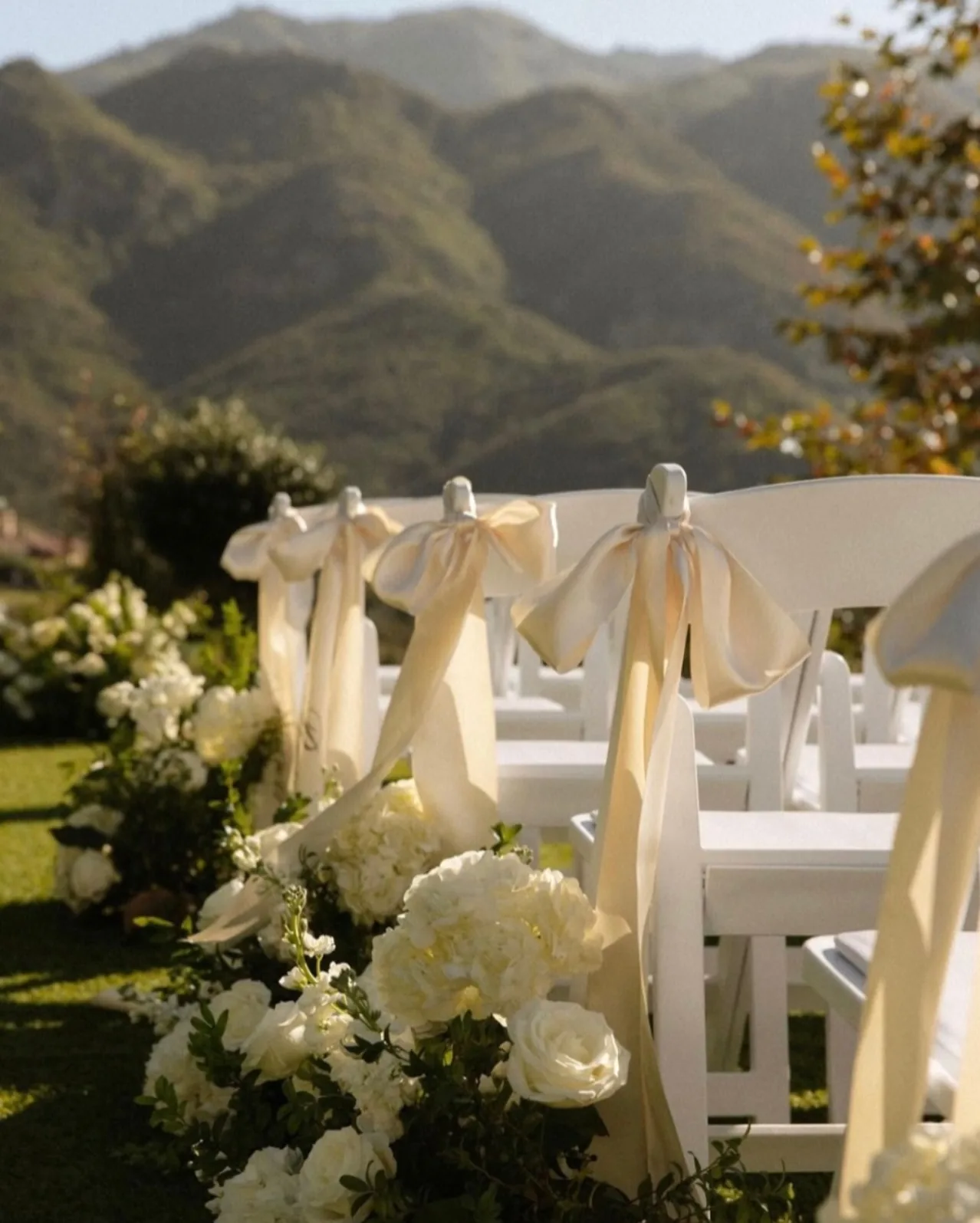 White folding chairs tied with ivory ribbons, with flowers at the aisle and green mountains behind